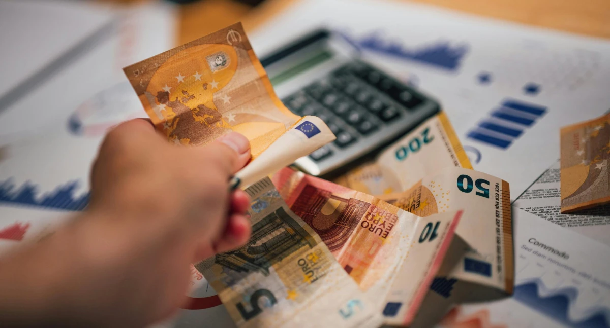 Hand holding euro banknotes over financial charts and calculator.